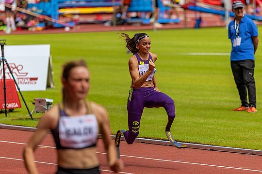 Eine Sportlerin mit Laufprothesen nimmt an einem Leichtathletikwettkampf teil; sie ist konzentriert und befindet sich mitten im Laufschritt. Im Vordergrund läuft eine andere Sportlerin vor ihr her. Im Hintergrund stehen Zuschauer und Offizielle neben der Laufbahn.