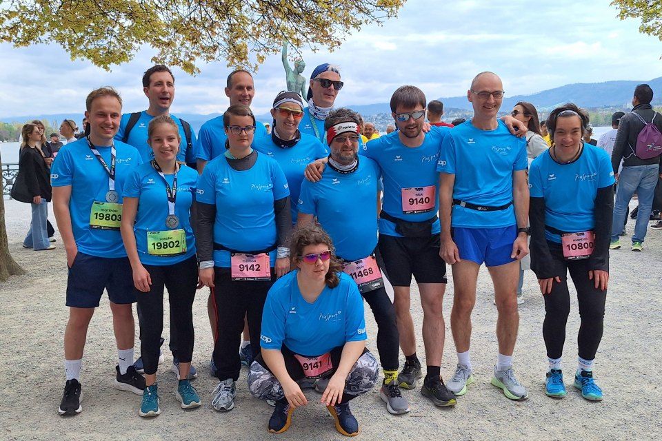 Eine Gruppe von dreizehn Personen in passenden blauen Shirts und Startnummern posiert für ein Foto im Freien an einem See, einige stehen und einige hocken und lächeln nach einem Rennen.