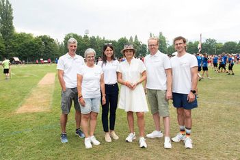 Sechs Erwachsene stehen nebeneinander auf einer Wiese, alle mit weißen Oberteilen und legeren Unterhosen. Im Hintergrund sind Bäume und Menschengruppen zu sehen, die auf eine Sport- oder Gemeinschaftsveranstaltung hindeuten.