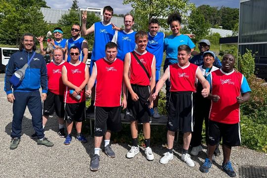 Eine Gruppe von Sportlern in roten und blauen Trikots posiert und lächelt im Freien, einige stehend und einige kniend, mit Bäumen und Gebäuden im Hintergrund an einem sonnigen Tag.