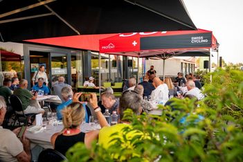 Eine Gruppe von Menschen sitzt an Tischen im Freien unter roten Swiss Cycling Zelten und hört einem Redner zu. Einige Leute machen Fotos, andere unterhalten sich, was wie eine organisierte Veranstaltung oder ein Treffen aussieht.