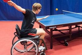 Eine Person im Rollstuhl spielt in einer Halle Tischtennis und bereitet sich darauf vor, den Ball mit einem roten Schläger zu schlagen. Der Tisch ist blau, und die Umgebung hat einen roten Boden und blaue Wände.