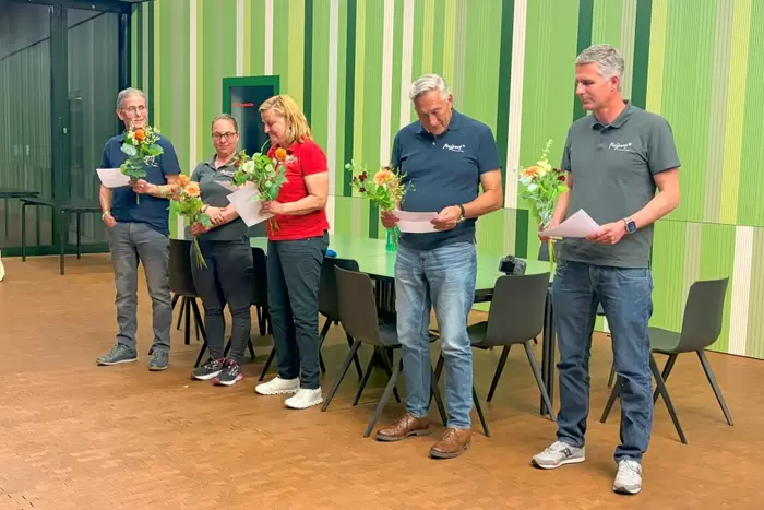 Fünf Personen stehen in einem Raum vor einer grün gestreiften Wand. Sie halten Papiere und Blumensträuße in den Händen. Dahinter steht ein Tisch mit Stühlen. Drei lesen oder schauen in Papiere, während zwei sich anlächeln.
