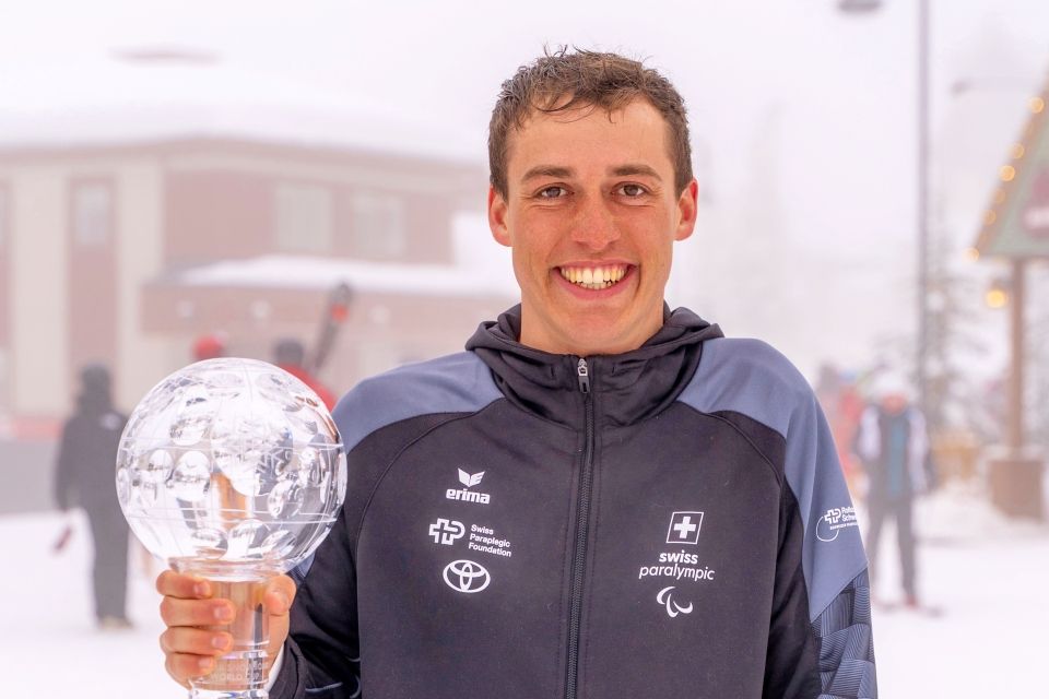 Ein lächelnder Athlet in einer Schweizer Paralympics-Jacke hält eine gläserne Trophäe. Um ihn herum fällt sanft Schnee, und im unscharfen Hintergrund sind ein verschneites Gebäude und Menschen zu sehen.