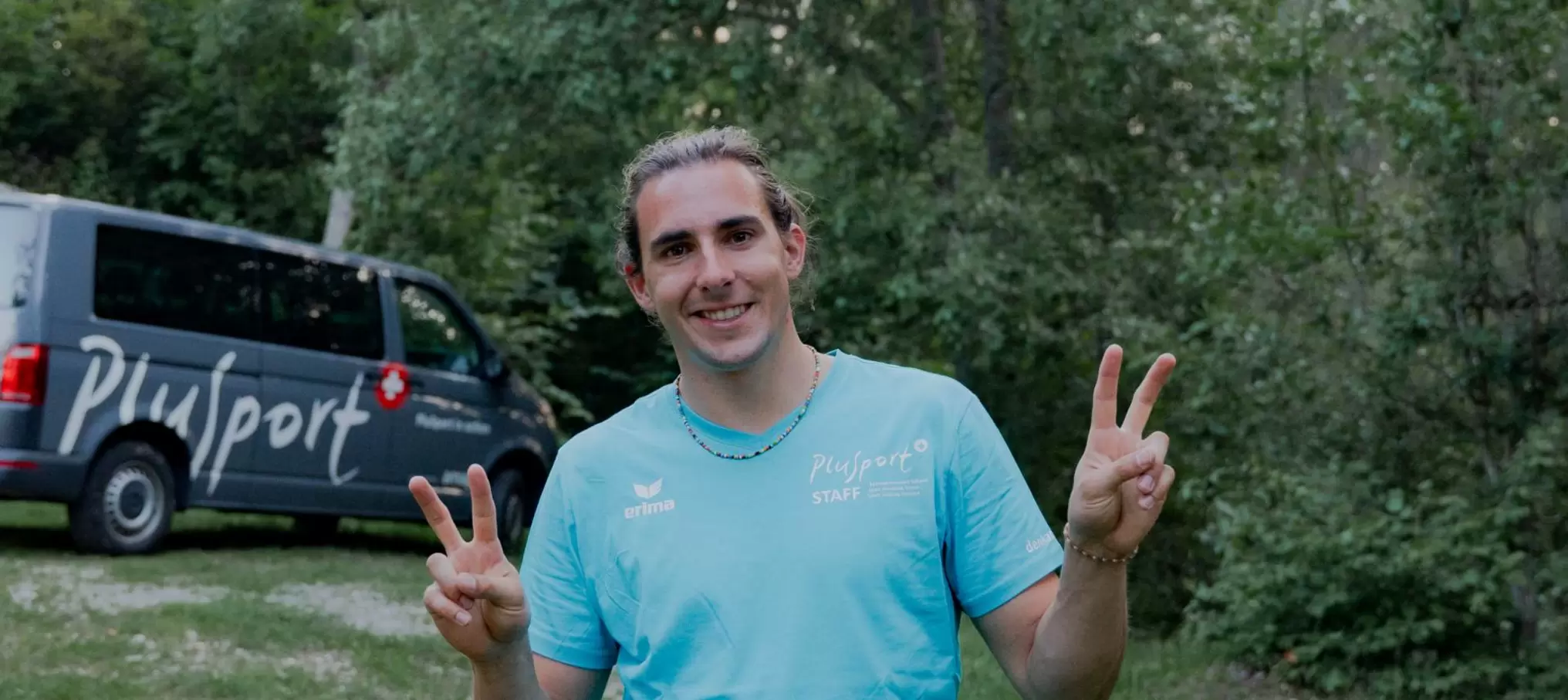 Eine Person mit langen Haaren, die ein hellblaues Hemd trägt, lächelt und formt mit beiden Händen das Peace-Zeichen. Im Hintergrund sind ein grauer Transporter mit der Aufschrift Pulsport und grüne Bäume zu sehen.