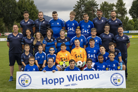 Eine Jugendfußballmannschaft und ihre Trainer, alle in blauen FC Widnau-Trikots, posieren auf einem Spielfeld und halten ein Banner mit der Aufschrift "Hopp Widnou" und dem Vereinslogo seit 1931. Einige Spieler halten Fußbälle in der Hand.