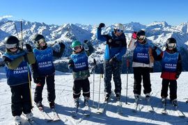 Fünf Kinder in blauen "Para Ski Training"-Westen und ein erwachsener Lehrer stehen mit Skiausrüstung auf einem verschneiten Berghang und heben die Arme, während im Hintergrund schneebedeckte Gipfel unter einem klaren blauen Himmel zu sehen sind.