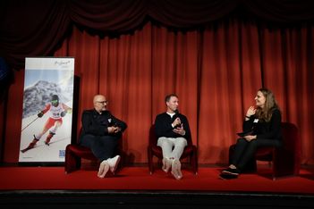 Drei Personen sitzen auf der Bühne vor roten Vorhängen und führen eine Diskussion. Links sitzen zwei Männer, einer hält ein Mikrofon, rechts sitzt eine Frau mit einem Notizblock. Im Hintergrund ist ein Skiplakat zu sehen.
