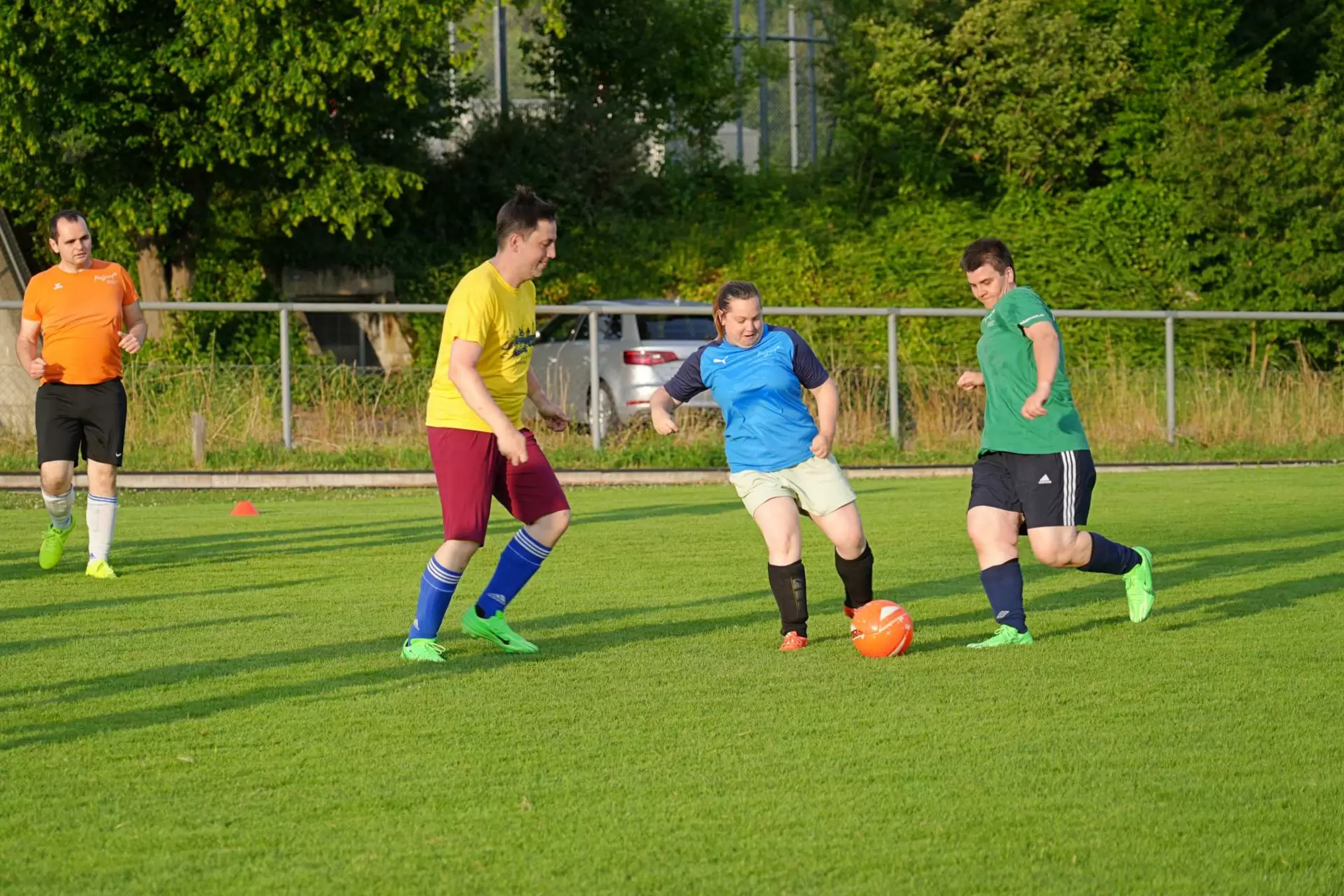 Vier Erwachsene spielen Fußball auf einer Wiese; einer in Grün versucht, einen orangefarbenen Ball zu schießen, während sich Spieler in blauen, gelben und orangefarbenen Hemden nähern. Im Hintergrund sind Bäume und ein geparktes Auto zu sehen.