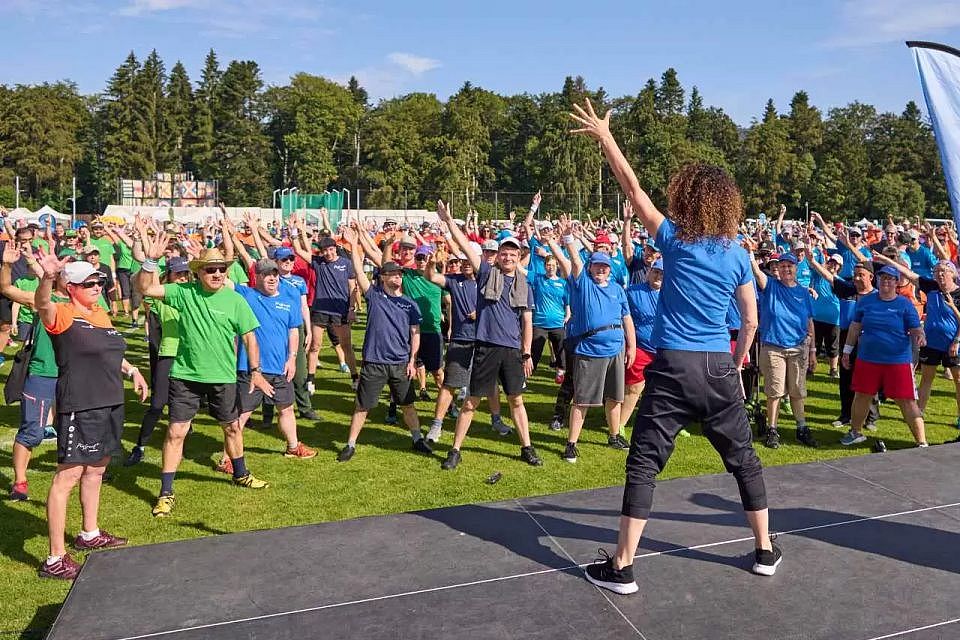 Eine große Gruppe von Menschen in farbenfroher Sportkleidung folgt einem Fitnesstrainer auf einer Bühne im Freien, alle heben einen Arm, auf einer Wiese mit Bäumen und einem klaren Himmel im Hintergrund.