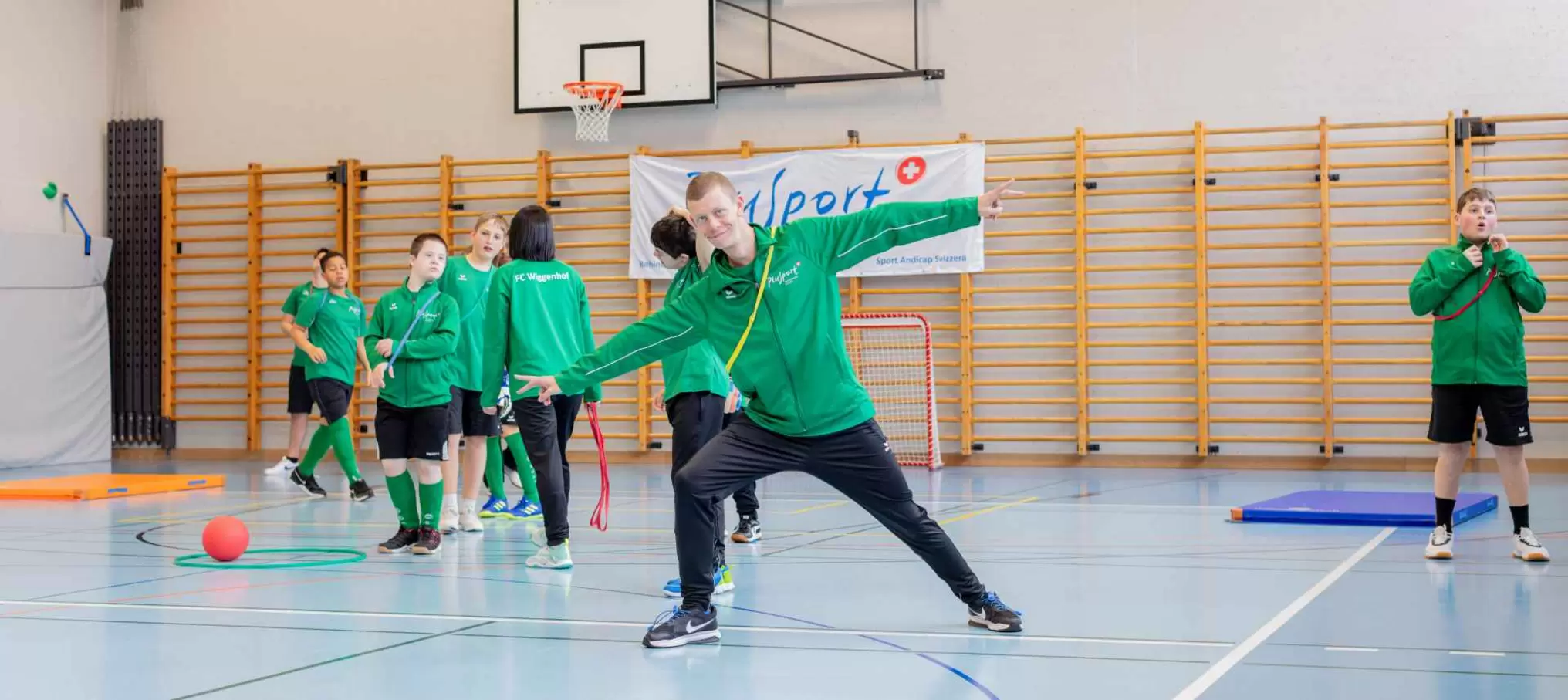 Eine Gruppe von Kindern in grünen Sportuniformen spielt und posiert in einer Turnhalle, im Hintergrund sind Sportgeräte und ein Basketballkorb zu sehen.