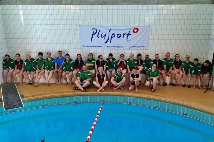 Eine Gruppe von Athleten und Trainern in grünen und blauen Uniformen sitzt und kniet neben einem Hallenbad und posiert für ein Gruppenfoto vor einem Plusport-Banner an einer gefliesten Wand.