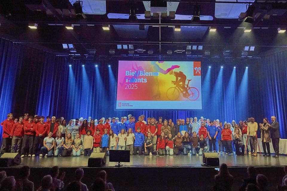Eine grosse Gruppe von Menschen posiert auf einer Bühne unter hellen Lichtern, während hinter ihnen ein buntes Banner von Biel/Bienne Talents 2025 mit einem Radfahrer auf einer Leinwand zu sehen ist. Im Vordergrund ist das Publikum zu sehen.