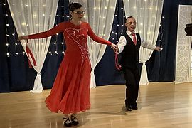 Zwei Menschen tanzen in einem Ballsaal. Die Frau trägt ein wallendes rotes Kleid mit weißen Mustern, und der Mann trägt einen schwarzen Anzug mit einer roten Krawatte. Weiße Vorhänge und Lichterketten schmücken den Hintergrund.