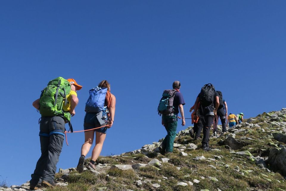 Eine Gruppe von Wanderern mit Rucksäcken erklimmt einen felsigen, grasbewachsenen Hügel unter einem strahlend blauen Himmel. Einige sind mit einem Seil verbunden, und alle gehen den Pfad hinauf.