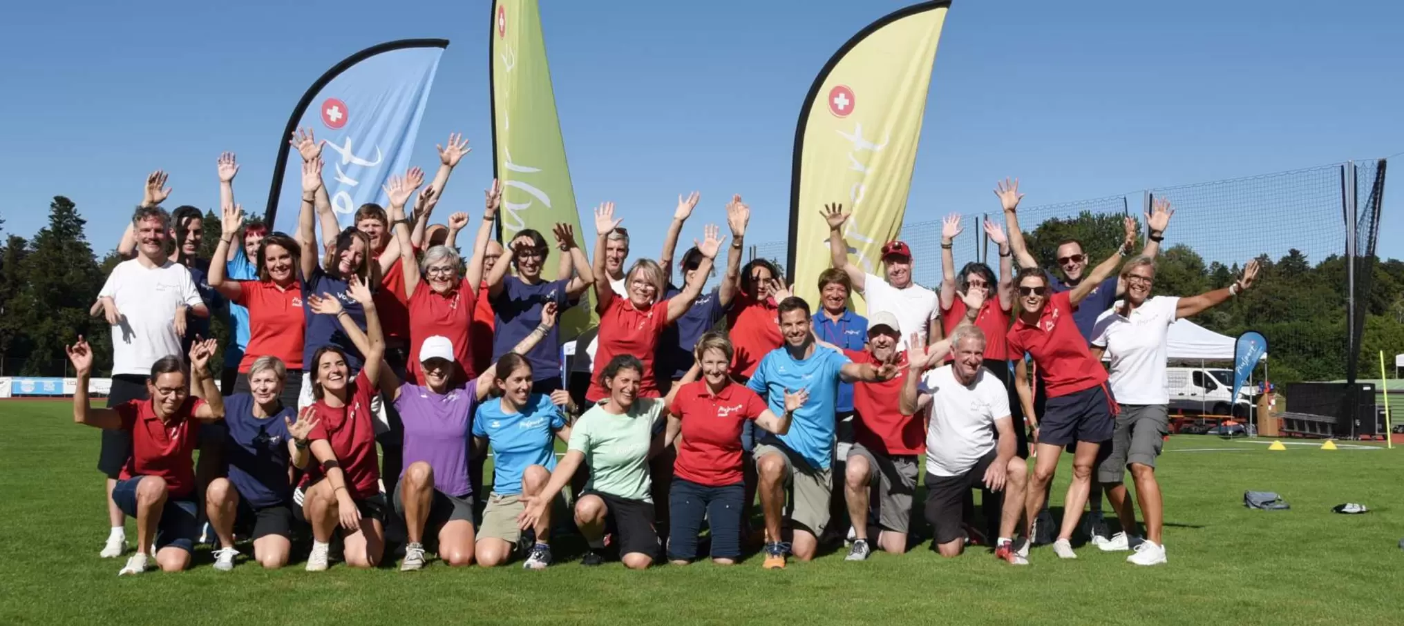 Eine große Gruppe von Menschen in Sportkleidung posiert und jubelt draußen auf einer Wiese, hinter ihnen wehen zwei bunte Fahnen, über ihnen erstreckt sich ein strahlend blauer Himmel.