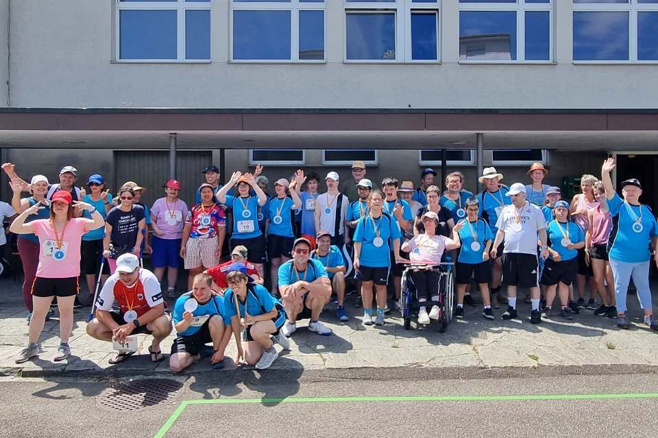 Eine große Gruppe von Menschen, viele mit Medaillen und blauen Hemden, posiert und winkt an einem sonnigen Tag vor einem Gebäude. Einige stehen, andere knien oder sitzen, lächeln und wirken glücklich nach einer Veranstaltung im Freien.