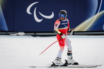 Ein paralympischer alpiner Skifahrer in Rennkleidung und Helm steht im Schnee und hält einen Skistock. Im Hintergrund ist das paralympische Symbol auf einer blauen Fahne zu sehen.