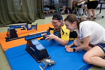 Zwei junge Frauen liegen auf Matten und zielen mit blauen Gewehren auf eine Zielscheibe. Die eine trägt ein gelbes Shirt und eine Mütze, während die andere ihr lächelnd Anweisungen gibt. Ein Wertungsgerät und weitere Ausrüstung befinden sich in der Nähe, im Hintergrund sind weitere Personen zu sehen.