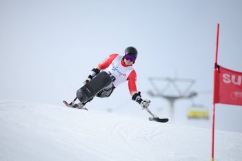 Ein alpiner Skifahrer in einem Sitzski fährt auf einer verschneiten Piste bergab. Er trägt einen Helm, eine Skibrille und eine weiß-rote Startnummer. Im Hintergrund sind eine rote Flagge und ein Skilift zu sehen.