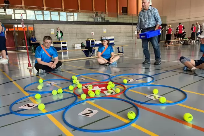 Menschen in blauen Hemden sitzen auf dem Boden einer Turnhalle um blumenförmig angeordnete Reifen, die mit Zahlen beschriftet sind. Tennisbälle sind in und um die Reifen herum verstreut. Ein älterer Mann steht in der Nähe und hält ein Tablett. Im Hintergrund sind weitere Personen zu sehen.