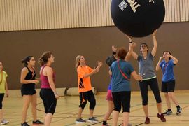 Eine Gruppe von Frauen in Sportkleidung spielt in einer Turnhalle ein Spiel mit einem großen schwarzen Ball mit der Aufschrift Kin-Ball. Einige Frauen greifen nach dem Ball, während andere zusehen oder sich darauf vorbereiten, mitzuspielen.
