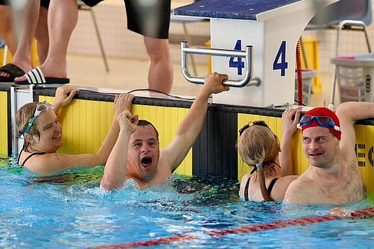Vier Schwimmer lächeln und feiern nach einem Rennen am Beckenrand, wobei ein Schwimmer vor Begeisterung die Faust hebt. Sie befinden sich in der Nähe einer Bahnmarkierung und des Startblocks mit der Nummer 4. Im Hintergrund stehen weitere Personen.