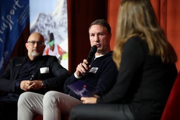 Drei Personen sitzen auf der Bühne auf Stühlen. Der Mann in der Mitte hält ein Mikrofon und spricht, während der Mann links und die Frau rechts zuhören. Im Hintergrund sind rote Vorhänge und ein Sporttransparent zu sehen.