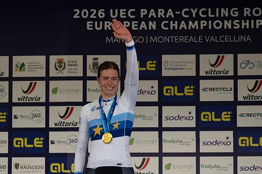 Ein lächelnder Radfahrer in einem weiß-blauen Siegertrikot und mit einer Goldmedaille um den Hals reckt auf dem Podium der UEC-Para-Cycling-Straßen-Europameisterschaft 2026 triumphierend den Arm in die Höhe. Im Hintergrund ist eine Sponsorenwand zu sehen.