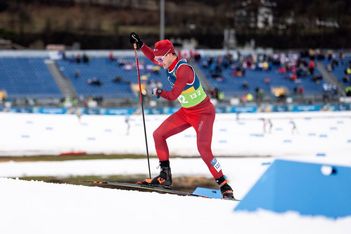 Ein Skilangläufer in roter Kleidung und mit Mütze fährt auf einer verschneiten Strecke mit Skistöcken bergauf. Im Hintergrund sind Zuschauer und blaue Absperrungen zu sehen, außerdem Schneeflecken und eine leere Tribüne.