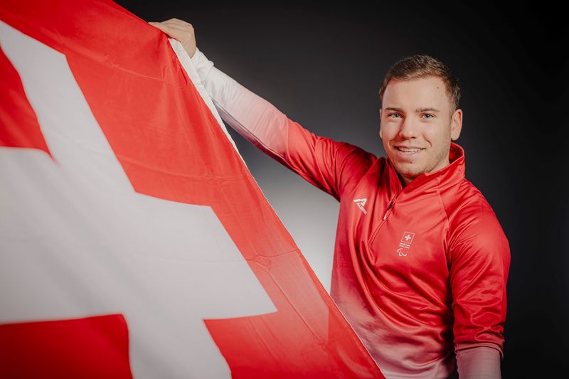 Eine Person in einer roten Jacke lächelt, während sie eine große Schweizer Flagge mit einem weißen Kreuz auf rotem Hintergrund vor einem dunklen Hintergrund hält.