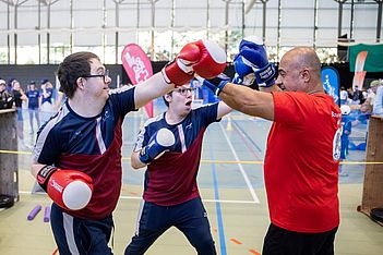 Zwei junge Männer mit Boxhandschuhen trainieren mit einem Trainer in einer Sporthalle. Der Trainer trägt ein rotes Shirt und blaue Handschuhe, während die jungen Männer aufeinander abgestimmte Outfits in Bordeauxrot und Blau tragen. Im Hintergrund sind weitere Personen und Transparente zu sehen.