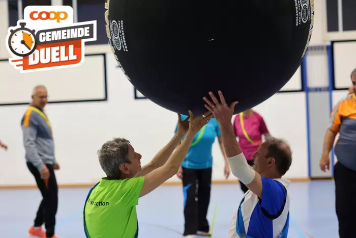 Zwei ältere Erwachsene in Sportkleidung heben in einer Turnhalle gemeinsam einen großen schwarzen Ball hoch, im Hintergrund sind weitere Personen und das Logo der Coop Gemeinde Duell zu sehen.