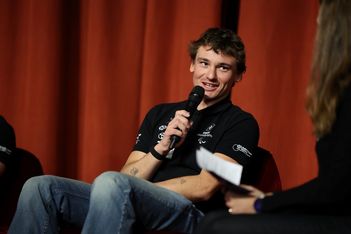 Ein junger Mann in schwarzem Hemd und Jeans sitzt da und lächelt, während er ein Mikrofon hält und während einer Veranstaltung spricht. Eine Person, die Papiere in der Hand hält, sitzt neben ihm. Im Hintergrund ist ein roter Vorhang zu sehen.