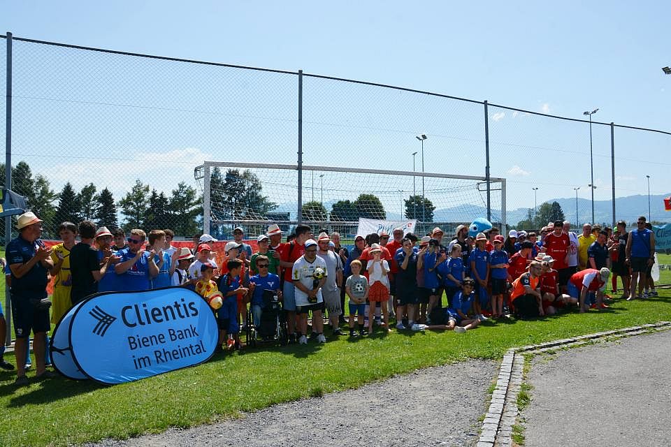 Eine große Gruppe von Menschen, darunter Kinder und Erwachsene, steht gemeinsam auf einem Fußballplatz in der Nähe eines Banners mit der Aufschrift „Clientis Biene Bank im Rheintal“. Es ist sonnig, und einige tragen Hüte und Sportkleidung.