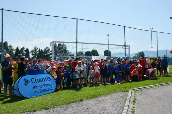 Eine große Gruppe von Menschen, darunter Kinder und Erwachsene, steht gemeinsam auf einem Fußballplatz in der Nähe eines Banners mit der Aufschrift „Clientis Biene Bank im Rheintal“. Es ist sonnig, und einige tragen Hüte und Sportkleidung.