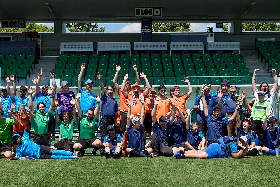 Eine große Gruppe unterschiedlicher Menschen in bunter Sportkleidung posiert fröhlich auf einem Fußballplatz, hebt die Arme und lächelt vor leeren grünen Stadionrängen unter strahlend blauem Himmel.