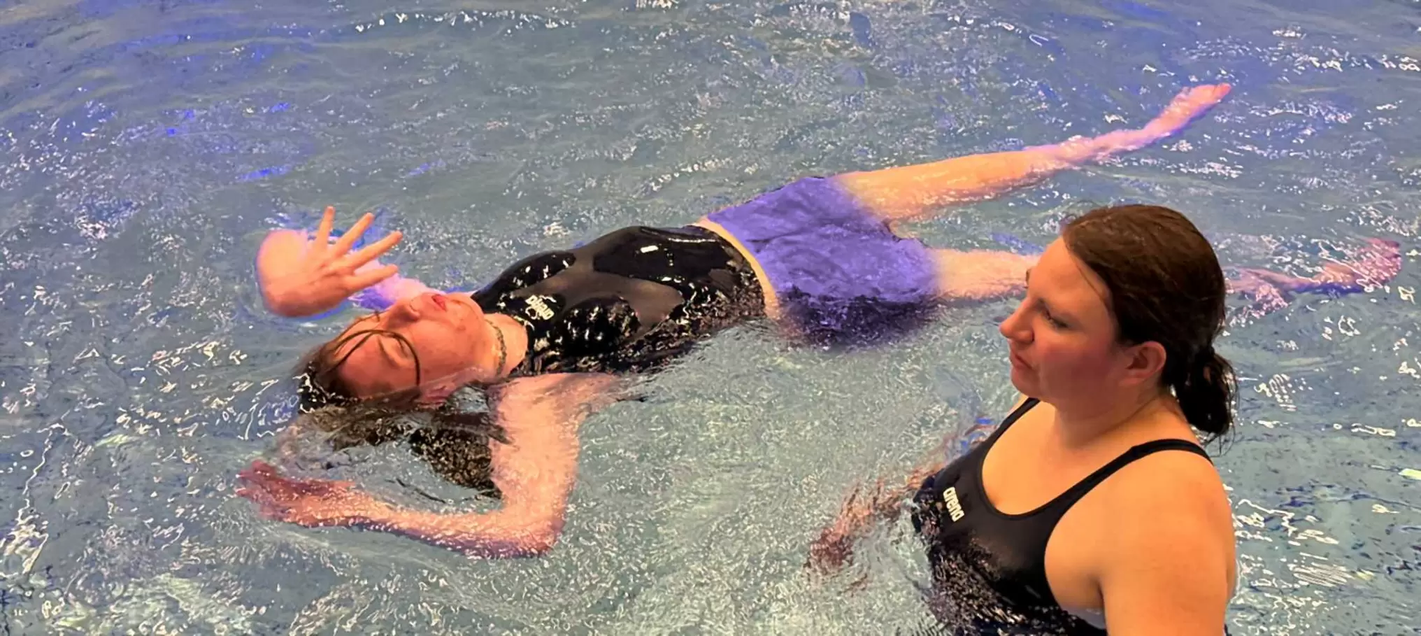 Zwei Frauen liegen in einem Schwimmbecken. Eine Frau schwimmt mit geschlossenen Augen und erhobenem Arm auf dem Rücken, während die andere daneben steht und aufmerksam zusieht.