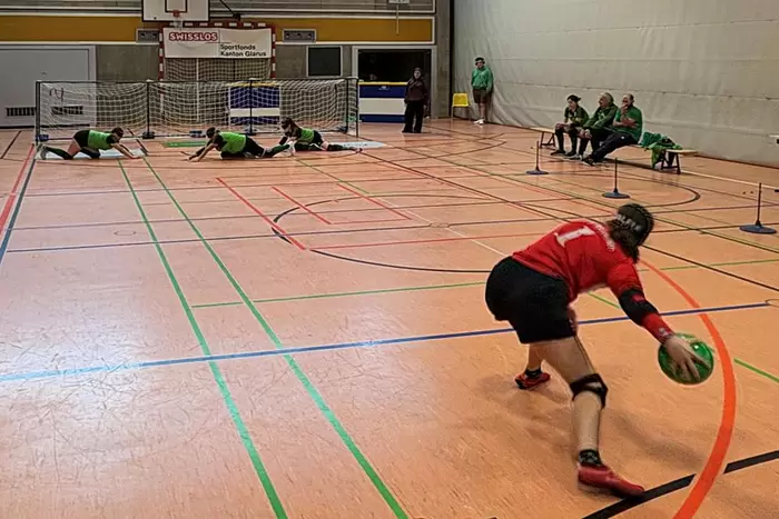 Ein Goalball-Spieler in einem roten Trikot bereitet sich auf den Wurf vor, während drei Spieler in grünen Trikots ein Tor auf einem Hallenplatz verteidigen. Zuschauer und Offizielle sitzen an der Seite.