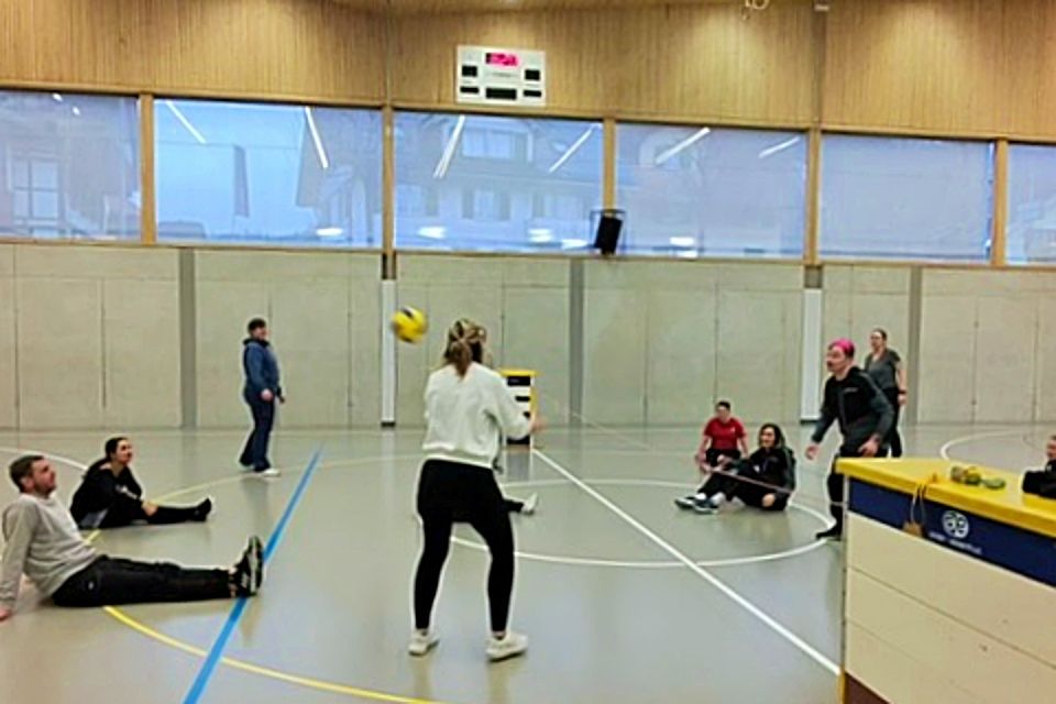 Eine Gruppe von Personen spielt Volleyball im Sitzen in einer Sporthalle. Die meisten Spieler sitzen auf dem Boden, während zwei in der Nähe des Netzes stehen und sich darauf vorbereiten, den Ball zu schlagen. Die Zuschauer schauen vom Rand aus zu.