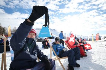 Eine Person in Winterkleidung lächelt und hält eine blaue Kuhglocke hoch. Sie sitzt auf einem Liegestuhl im Schnee, neben anderen, die in Decken mit Schweizer Fahne eingewickelt sind, mit Bergen und Fahnen im Hintergrund unter einem blauen Himmel.