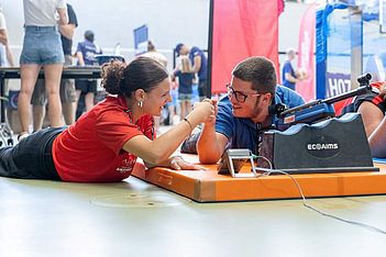 Zwei Personen, die auf Matten liegen, bestreiten ein Armdrücken und lächeln einander während einer Veranstaltung in der Halle an. Im Hintergrund sind Menschen und Stände zu sehen.