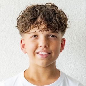 Ein lächelnder Junge mit lockigem braunem Haar und einem weißen T-Shirt steht vor einer weißen strukturierten Wand und blickt leicht nach oben.