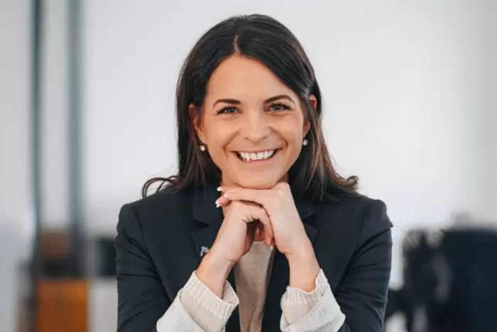 Eine Frau mit langen dunklen Haaren lächelt warm und stützt ihr Kinn auf ihre Hände. Sie trägt einen dunklen Blazer und eine helle Bluse und sitzt in einer hellen, modernen Inneneinrichtung.