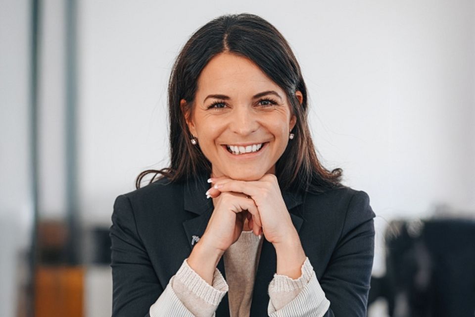 Eine Frau mit langen dunklen Haaren lächelt warm und stützt ihr Kinn auf ihre Hände. Sie trägt einen dunklen Blazer und eine helle Bluse und sitzt in einer hellen, modernen Inneneinrichtung.