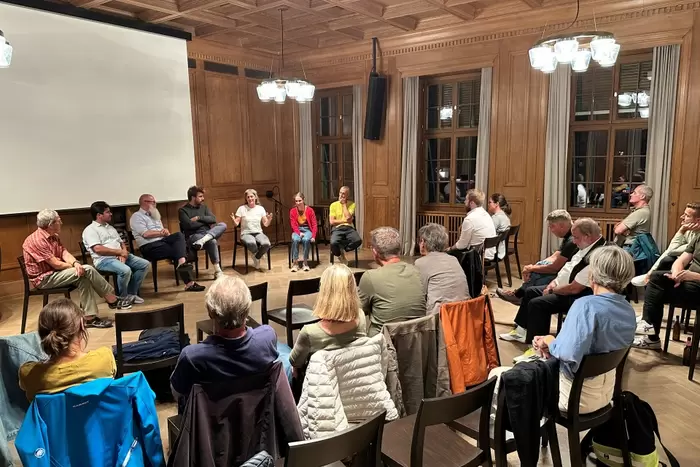 Eine Gruppe von Menschen sitzt im Kreis in einem holzgetäfelten Raum; sieben Personen an der Vorderseite leiten eine Diskussion, während die anderen aufmerksam zuhören.