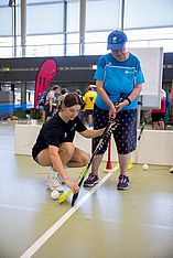 Eine junge Frau kniet sich hin, um einer älteren Frau zu helfen, die einen Floorballschläger in der Hand hält, und hilft ihr dabei, auf einem Hallenplatz auf den Ball zu zielen, während im Hintergrund andere Personen und Sportgeräte zu sehen sind.