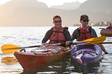 Zwei Menschen in roten Schwimmwesten und mit Sonnenbrillen sitzen lächelnd in roten und violetten Kajaks auf einem ruhigen See, im Hintergrund sind Berge und Sonnenschein zu sehen.