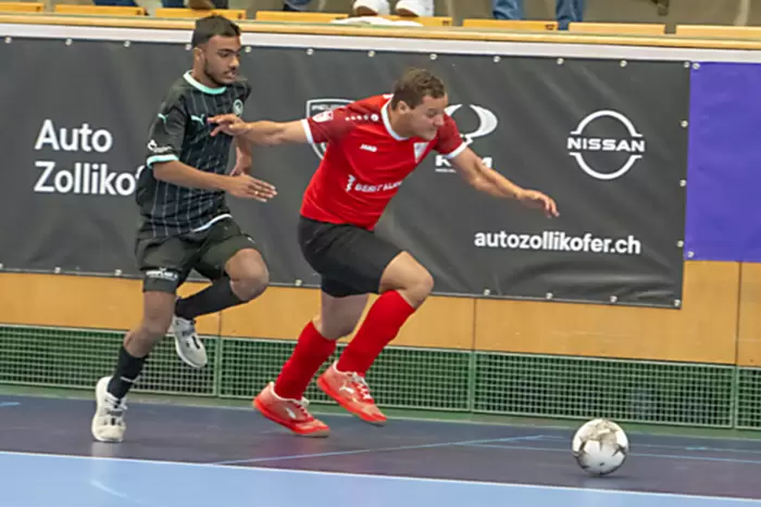 Zwei Männer spielen Hallenfußball. Einer in schwarzer Uniform jagt einen anderen in roter Uniform, während sie auf einen weißen Ball zurennen. Sie stehen auf einem blauen Boden, im Hintergrund sind Werbebanner zu sehen.