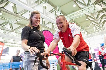 Ein lächelnder Mann in Sportkleidung fährt drinnen auf einem Heimtrainer, während eine Frau neben ihm ihn anfeuert und unterstützt. Sie befinden sich in einem hellen, geräumigen Saal, im Hintergrund sind Menschen und Transparente zu sehen.